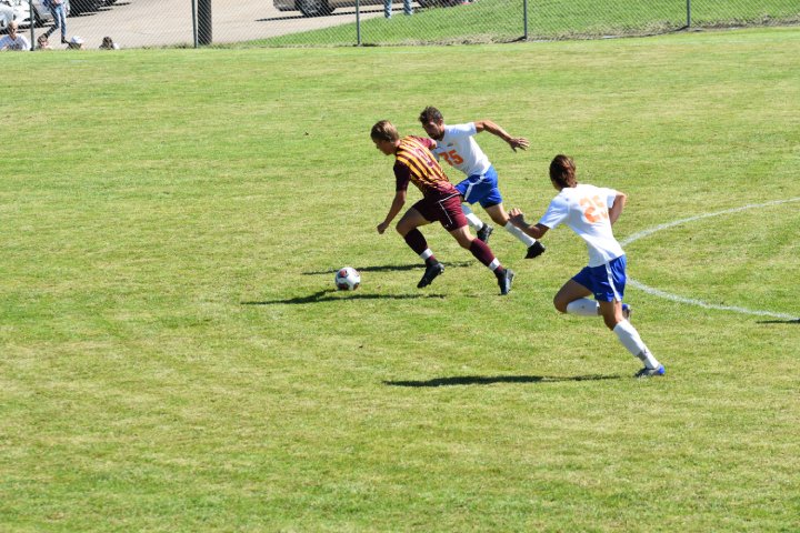 A soccer player in a maroon and gold jersey dribbles the ball down the field while being pursued by two opposing players in white jerseys with blue shorts. The action takes place on a grassy field on a sunny day, with part of a chain-link fence and some spectators visible in the background.